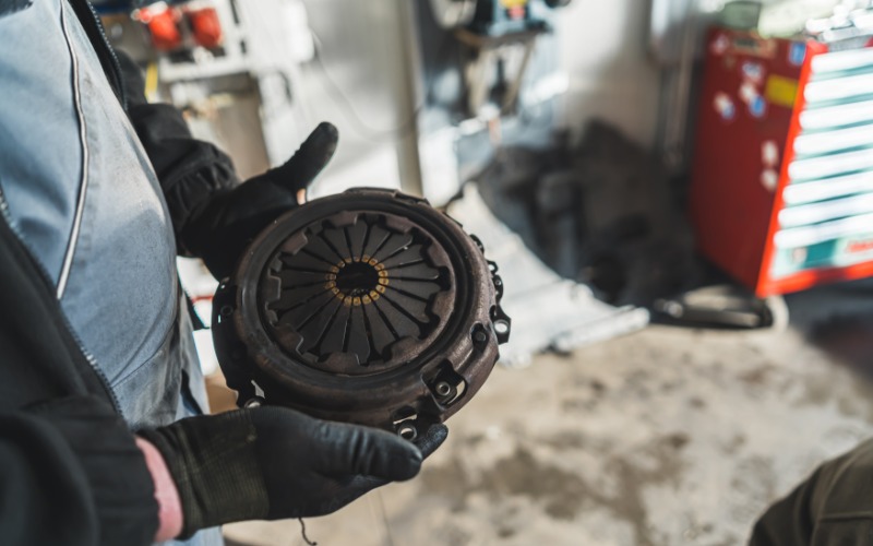 A close-up of a man with black gloves holding onto an old, used, and broken clutch in an auto repair shop.