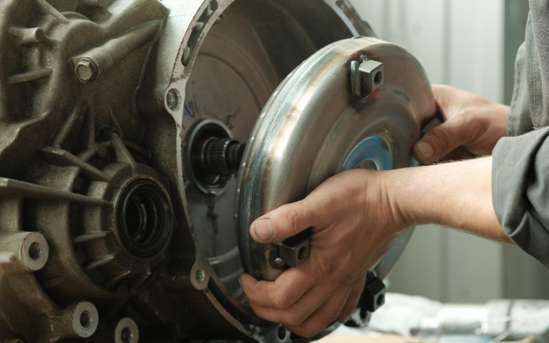 A close-up of a person's hands installing a large, round disc component into the transmission of a car.