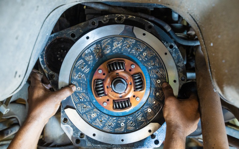 A close-up of two hands reaching into a vehicle's transmission to install a new clutch system, including the flywheel.