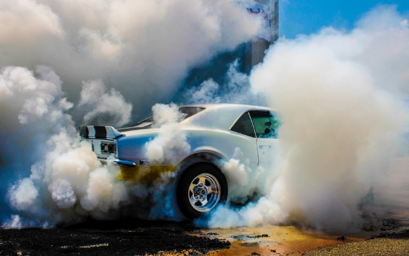 A rear view of a white classic muscle car with a spoiler and black racing stripes, creating smoke with a burnout.