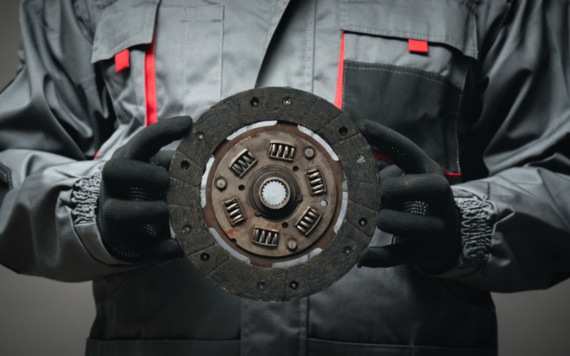 A close-up of the torso of a man with black gloves and a grey work jacket holding up an old car clutch disc.