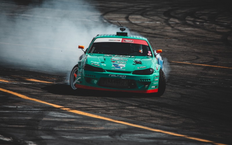 A teal sports car with numerous sponsor stickers drifting around the corner of a racetrack and generating smoke.