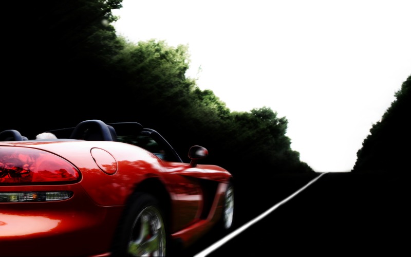 A rear view of a red, modern sports car with an open top and two doors driving on an empty road surrounded by trees.