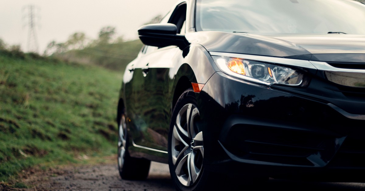 The front and side profile of a black, modern, compact sedan on a dirt road with green hills behind it.