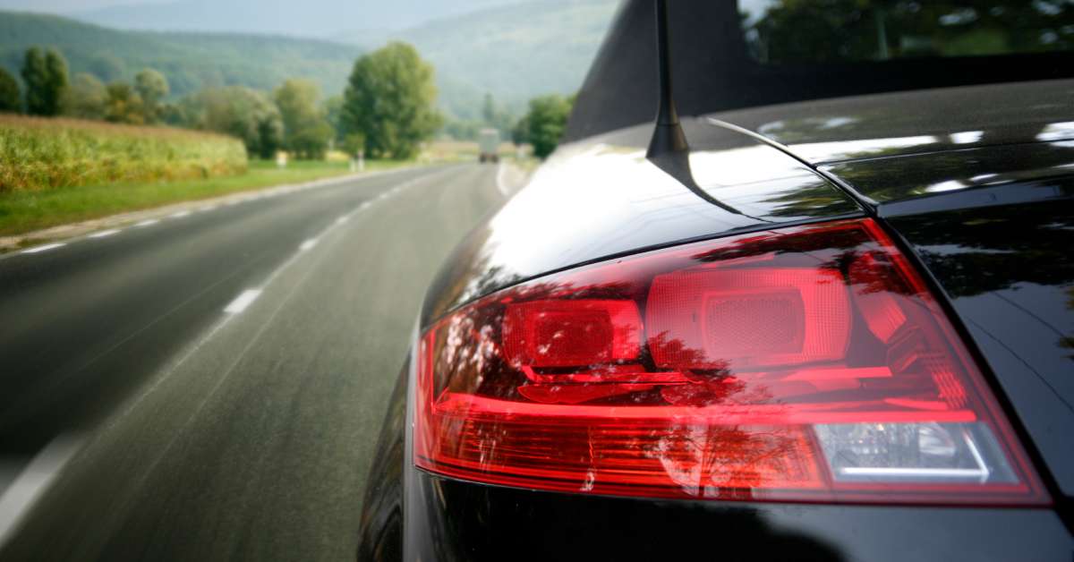 The red rear headlight and back end of a black Audi as it drives down a highway along a vibrant countryside.