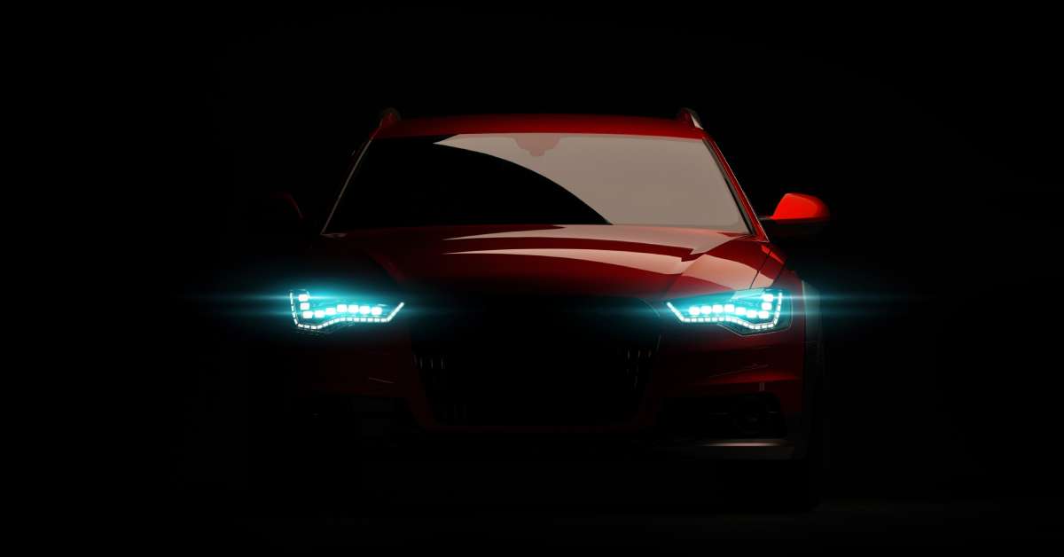 The front end of a red Audi with shadows covering most of the front end and the headlights activated.