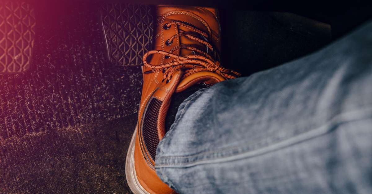 A driver, wearing brown shoes and jeans, presses on the gas pedal. The gas, brake, and clutch pedals are visible.