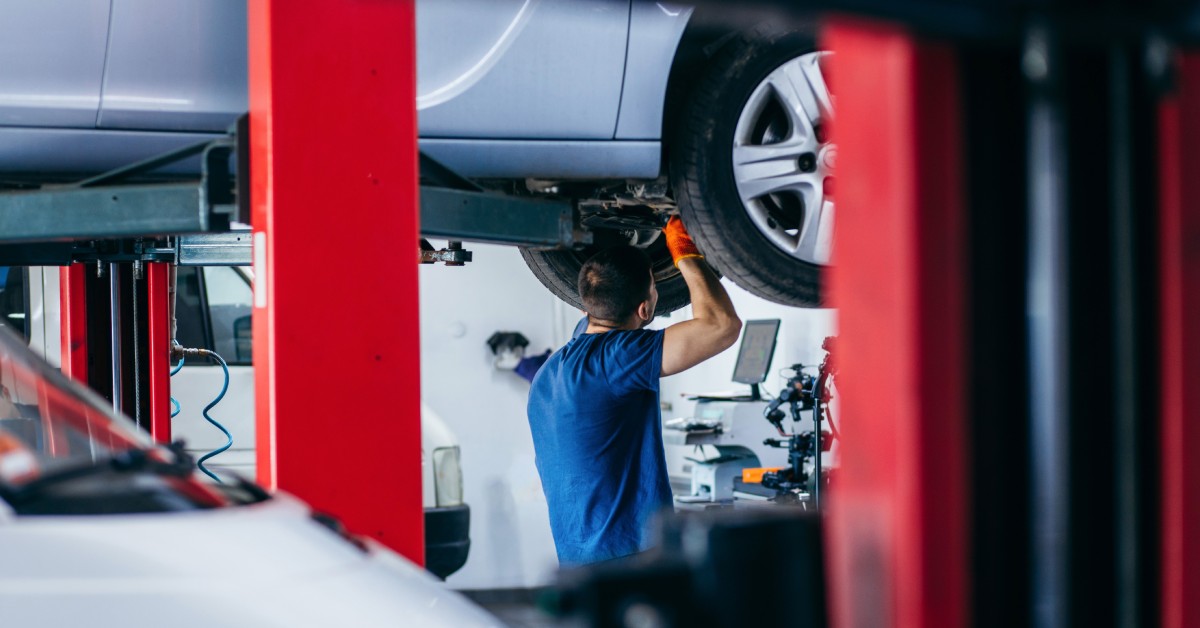 A male mechanic is working on the suspension and undercarriage of a car on a vehicle lift in a garage.