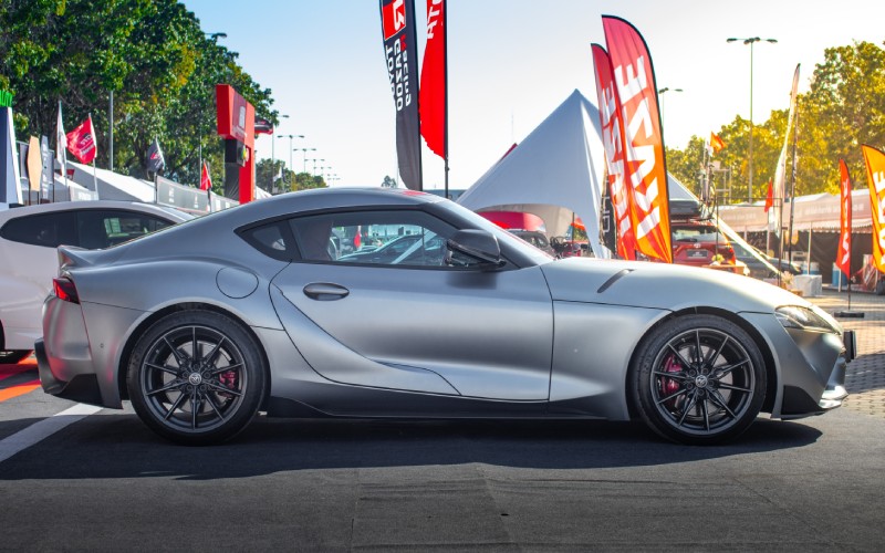 A silver Toyota Supra is on display at an auto convention and surrounded by display flags and tents.