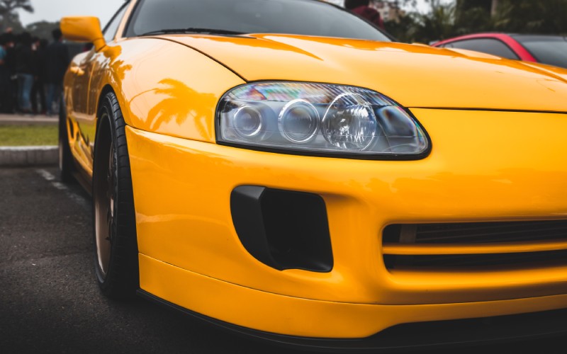 A yellow 2018 Toyota Supra is stopped in a parking lot. The focus is on the front bumper and headlight.