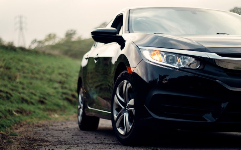 A front and side view of a black Honda sedan with lights activated on a dirt road with a grassy hill behind it.