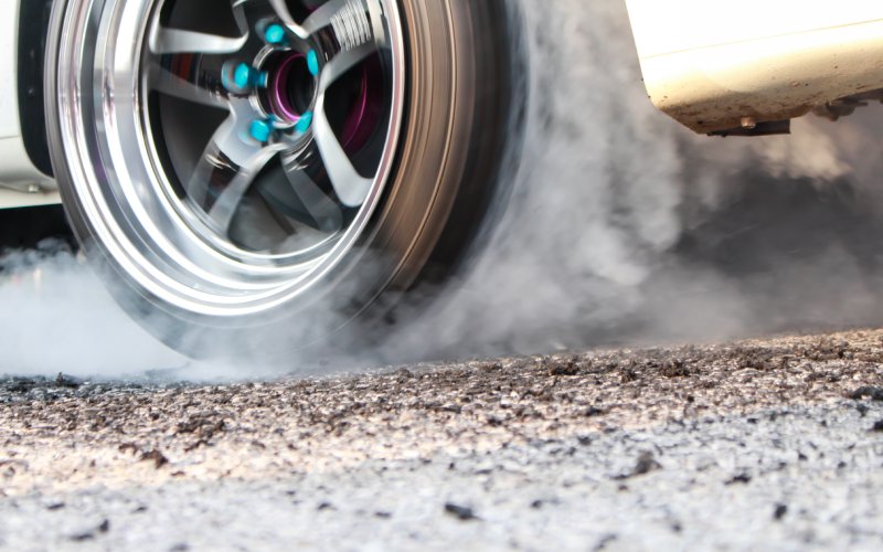 A close-up of the tire and wheel of a drag racing car starting on the pavement and generating white smoke.