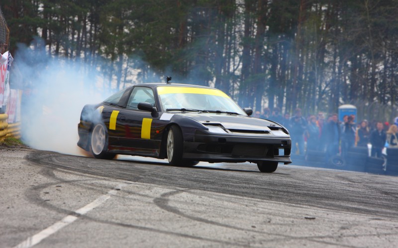 A black sports car with yellow accents drifts around the corner of a racetrack, producing smoke from the rear tires.