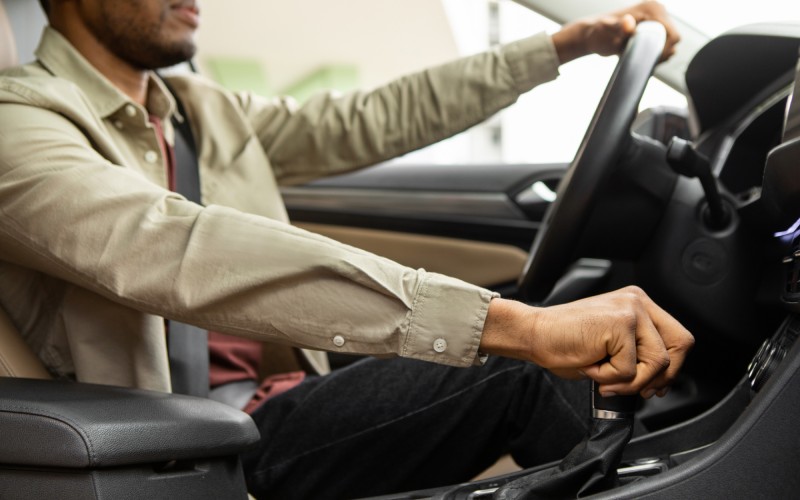 A side view of a driver in a tan dress shift holding the stick shift with one hand and the steering wheel with the other.