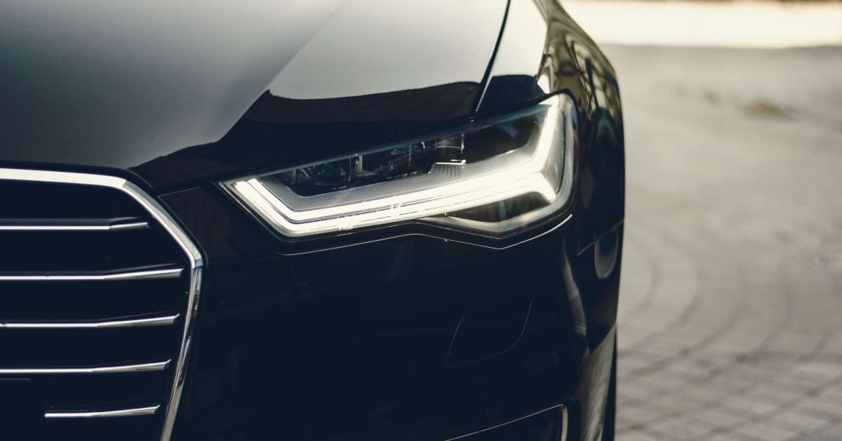 An individual front headlight and part of the chrome front end grille of a black Audi. The headlights are on.