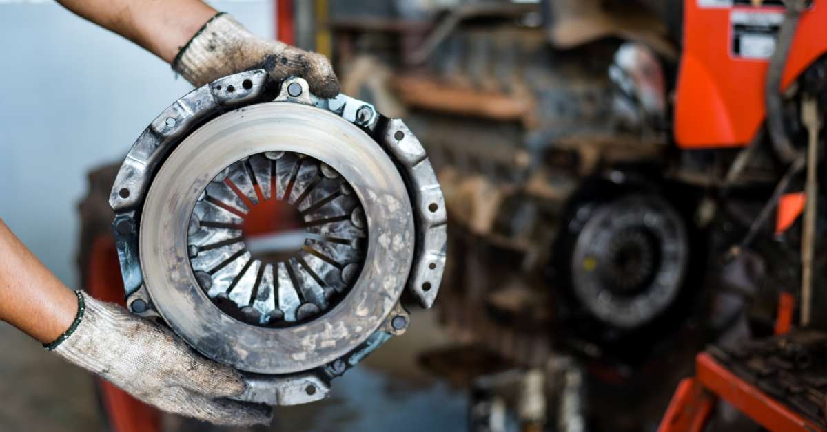 A mechanic wearing white work gloves holds up a dirty and used automotive clutch disc in a mechanic's garage.