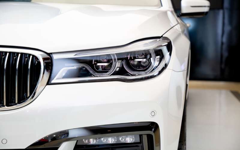 The front headlight, bumper, and grille of a white sedan. The car is parked on display in a showroom.